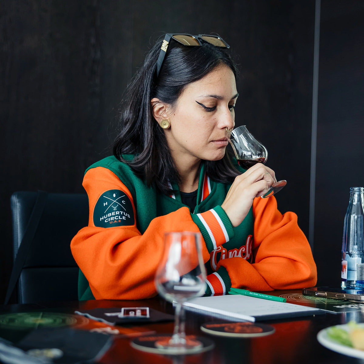 Person seated at a table holding a tasting glass close to their nose, with a notebook, pen, and additional glasses on the table, in an indoor training or tasting setting.