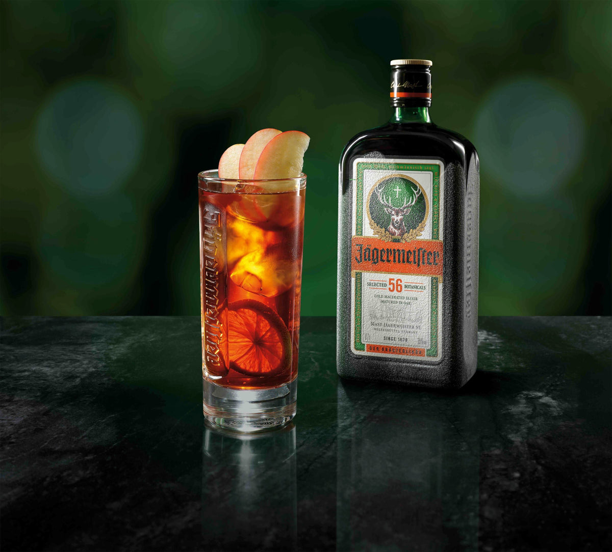 A bottle of Jägermeister stands next to a highball glass filled with an amber cocktail, garnished with apple slices and a lemon wheel, on a dark reflective surface with a blurred green background.