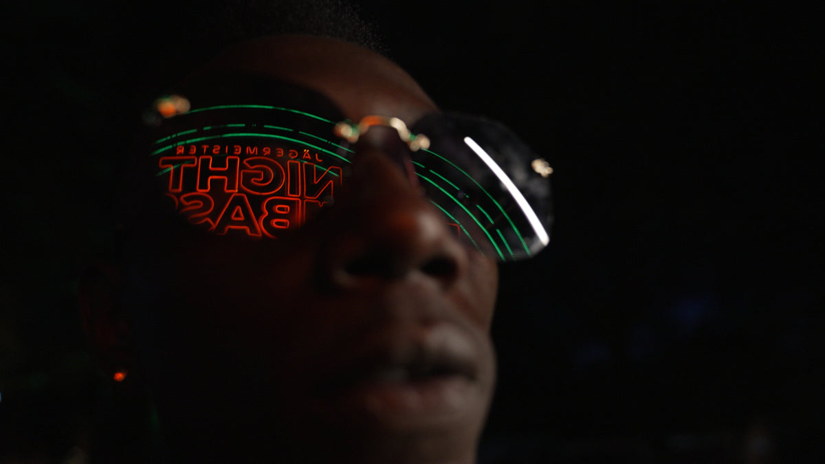 A close-up of a person wearing futuristic dark sunglasses with glowing green circular patterns and red mirrored lenses reflecting the word 'night embassy'.
