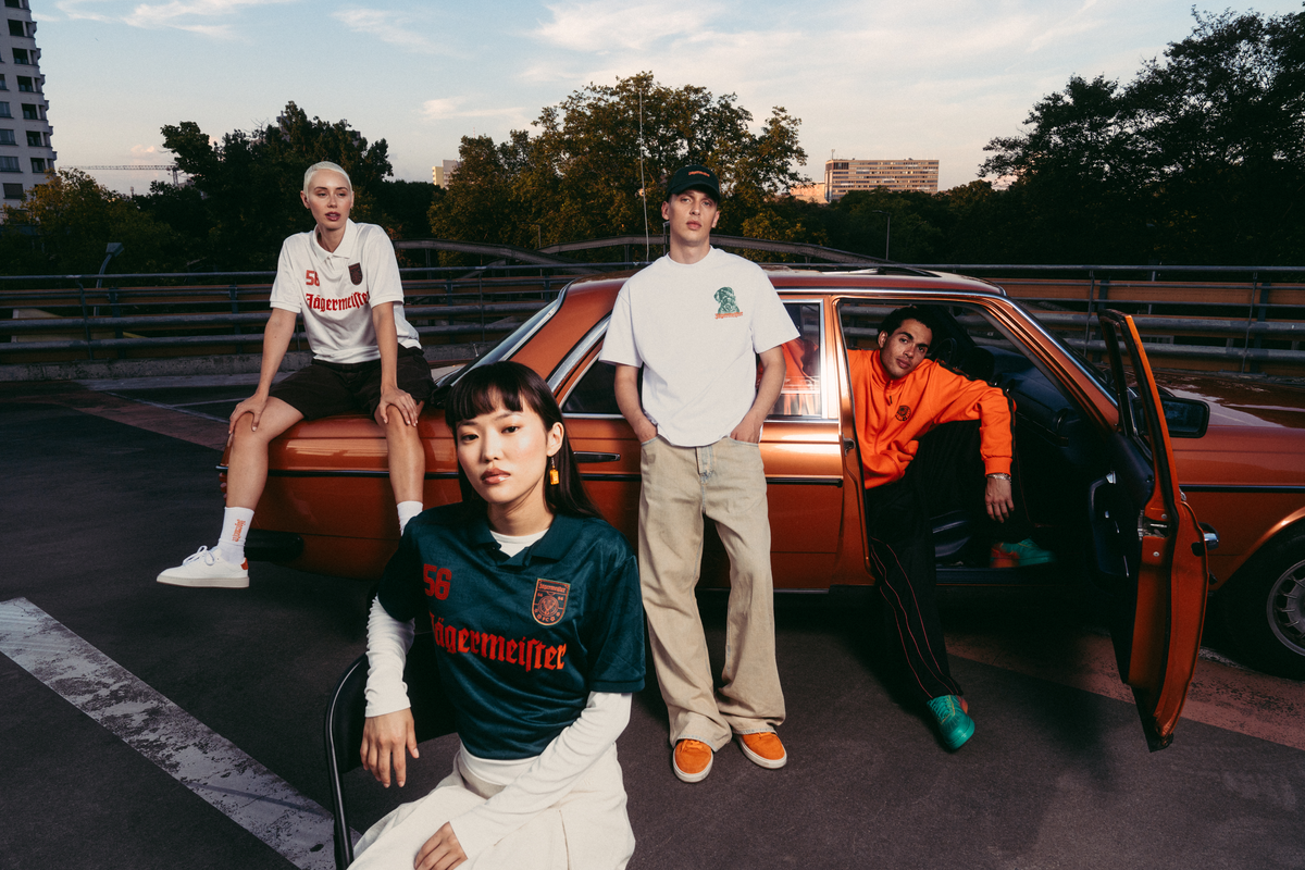 Four models posing with a vintage orange car on an outdoor bridge at dusk. One model sits on the car hood wearing a white Jägermeister jersey with a orange graphic, another stands in the middle wearing a white Jägermeister Waldi t-shirt and beige pants, a third model sits in the driver's seat wearing an orange Jägermeister hoodie, and a fourth model sits in the foreground wearing a dark green Jägermeister jersey.