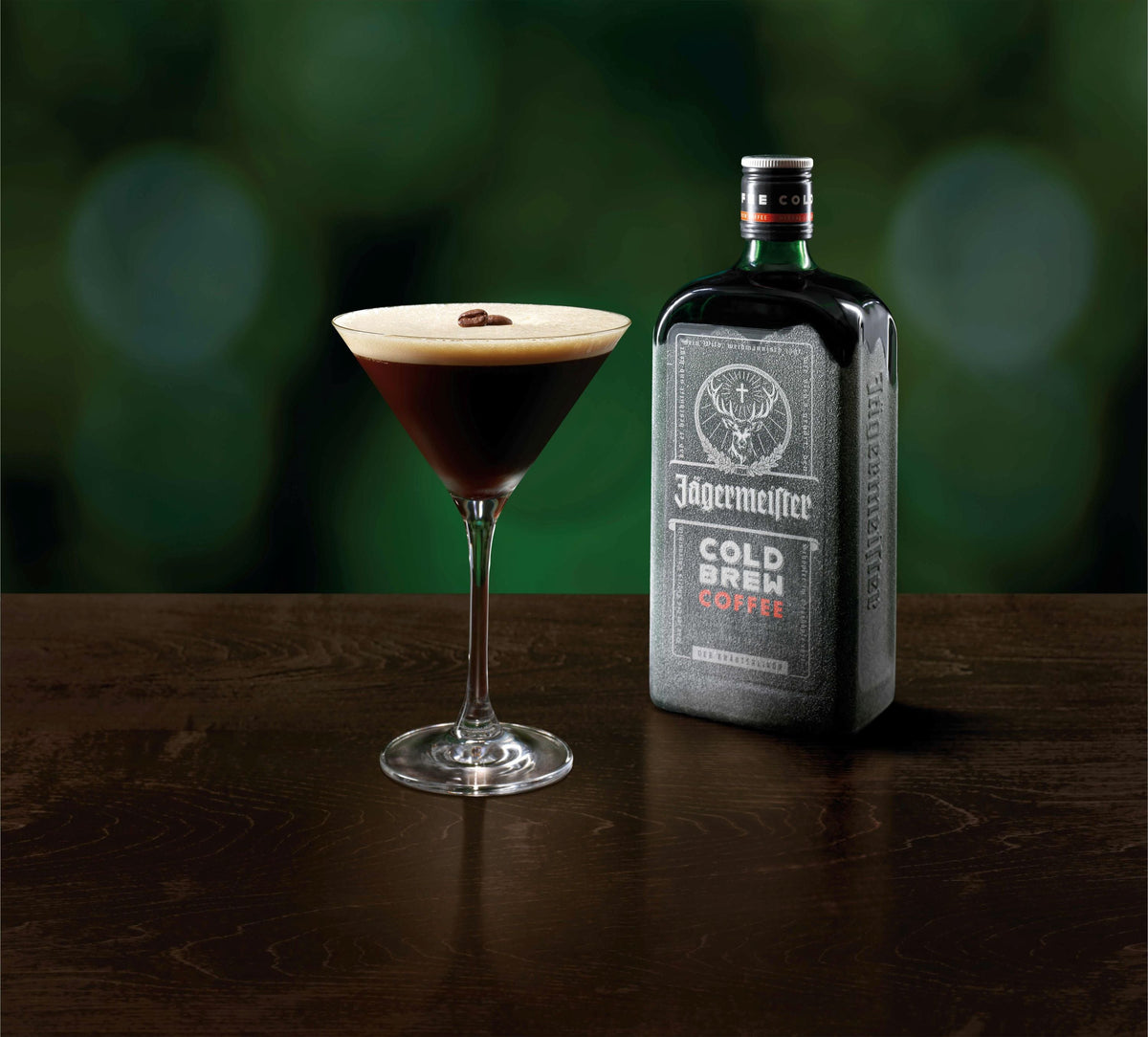 A Jägermeister Cold Brew Coffee cocktail in a coupe glass with a creamy foam top and a coffee bean garnish, displayed next to a bottle of Jägermeister Cold Brew Coffee on a dark wooden surface against a blurred green background.