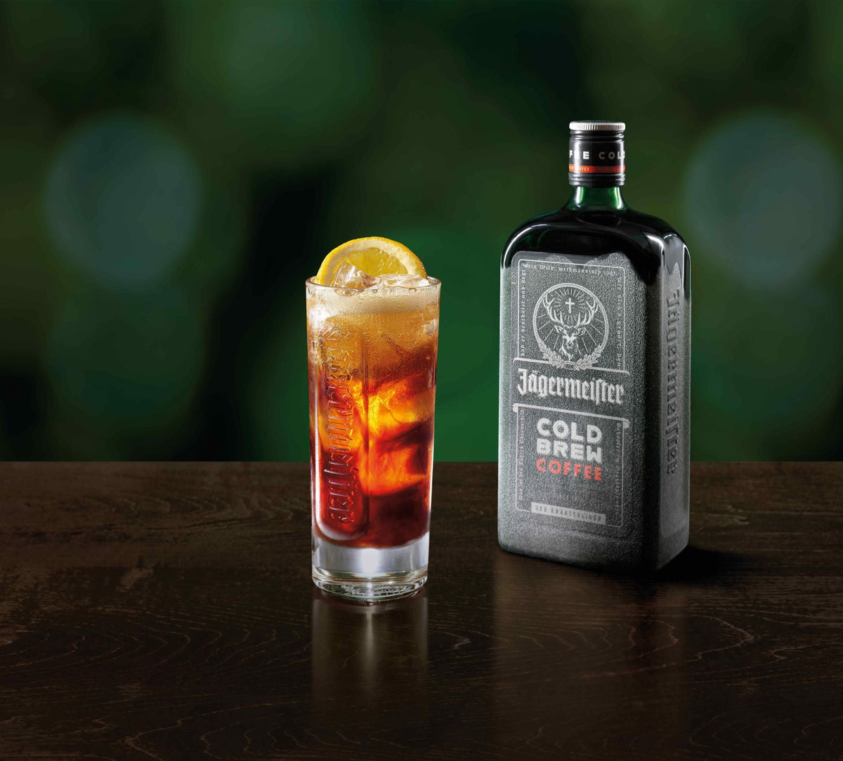 A bottle of Jagermeister Cold Brew Coffee stands next to a tall glass of iced coffee with a lemon slice garnish on a dark wooden surface against a blurred green background.