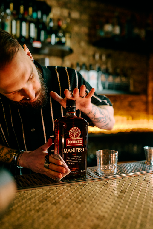 A bartender with a beard and tattoos leans over a bar counter, holding a bottle of Jägermeister Manifest. The bottle is positioned next to a clear rocks glass and a metal jigger, with a warm, blurred bar background.