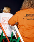 Două persoane așezate pe scaune verzi și albe, văzute din spate, purtând tricouri Jägermeisters. Un tricou este alb, cu un imprimeu portocaliu al etichetei, iar celălalt este portocaliu aprins, cu adresa sediului Jägermeister scrisă cu negru.