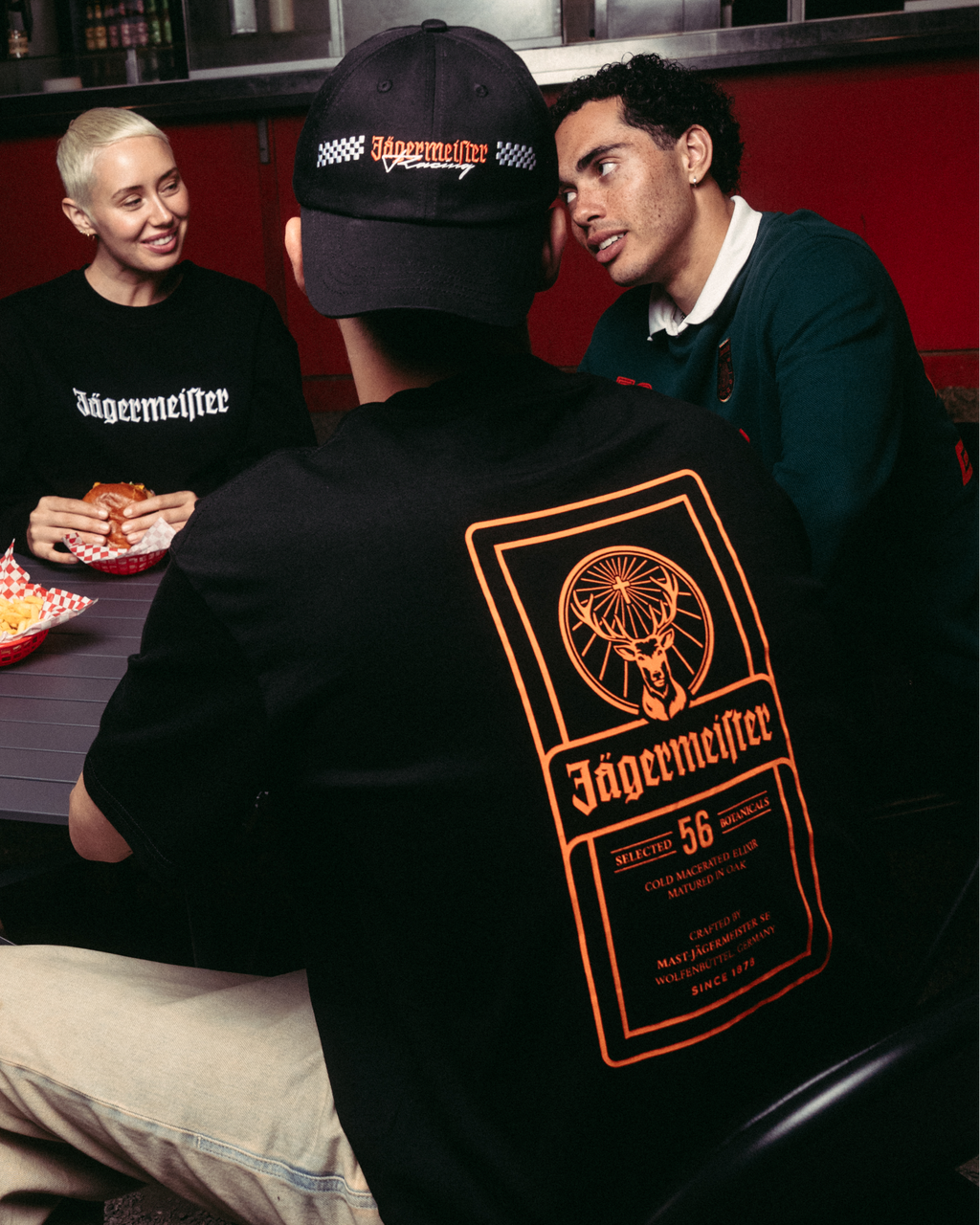 Patru persoane adunate în jurul unei mese la un stand de mâncare, cu accentul principal pe o persoană care poartă un tricou negru Jägermeister cu un grafic mare portocaliu pe spate, pantaloni khaki și o șapcă neagră.