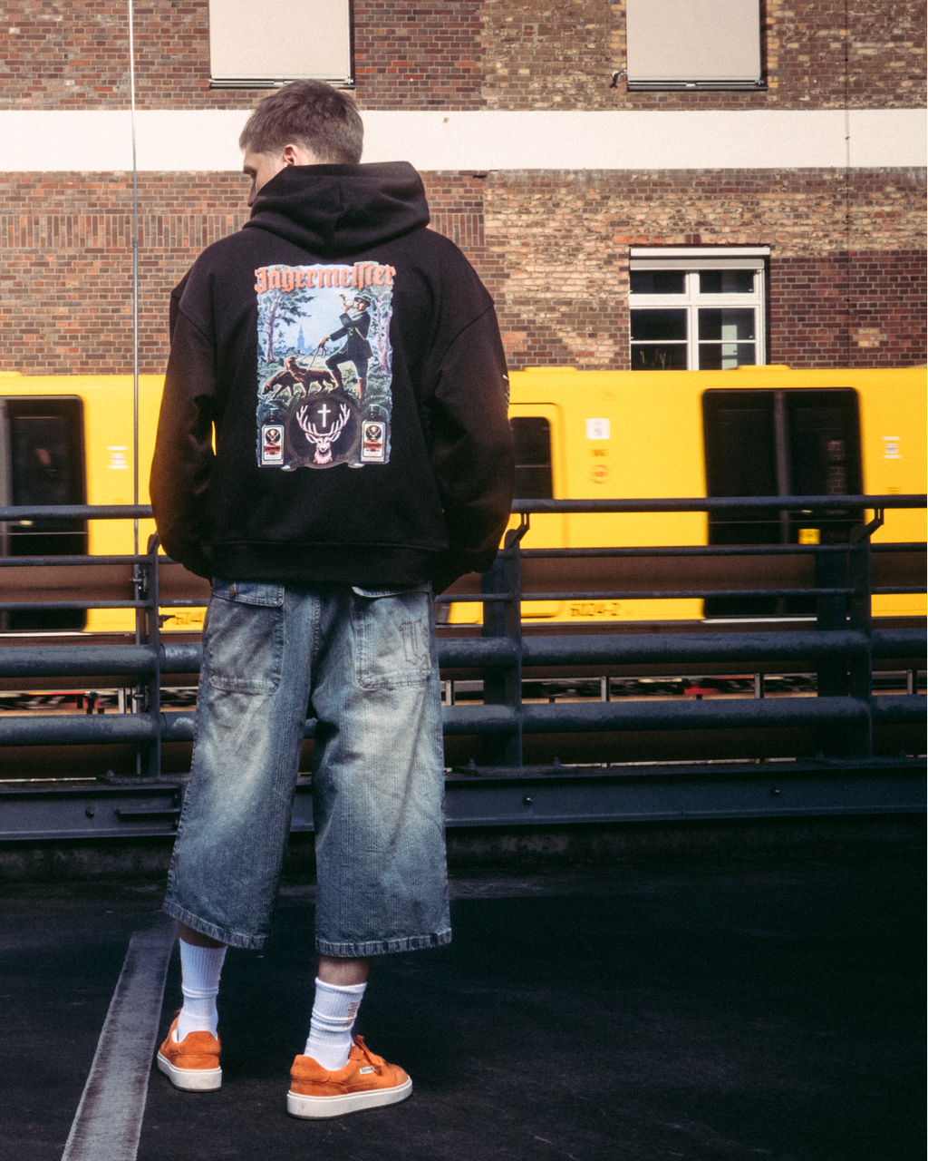 Un model purtând articole vestimentare streetwear, inclusiv un hanorac negru cu imprimeu mare pe spate Jägermeister tradition graphic, stând pe o suprafață pavată întunecată, în fața unei clădiri din cărămidă și a unui tren galben.
