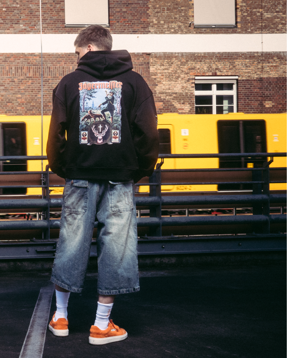 Un model purtând articole vestimentare streetwear, inclusiv un hanorac negru cu imprimeu mare pe spate Jägermeister tradition graphic, stând pe o suprafață pavată întunecată, în fața unei clădiri din cărămidă și a unui tren galben.