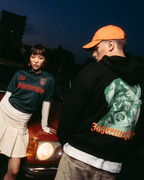 Două modele pozând în fața unei mașini vintage roșii, la amurg. Un model poartă un jersey verde închis Jägermeister cu o fustă albă plisată, iar celălalt poartă un hanorac negru Jägermeister Waldi și o șapcă portocalie Jägermeister Dad cap.
