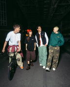 Patru persoane merg pe jos și pe bicicletă pe o stradă întunecată, noaptea, sub o linie de tren supraterană, purtând haine streetwear Jägermeister în diferite culori.