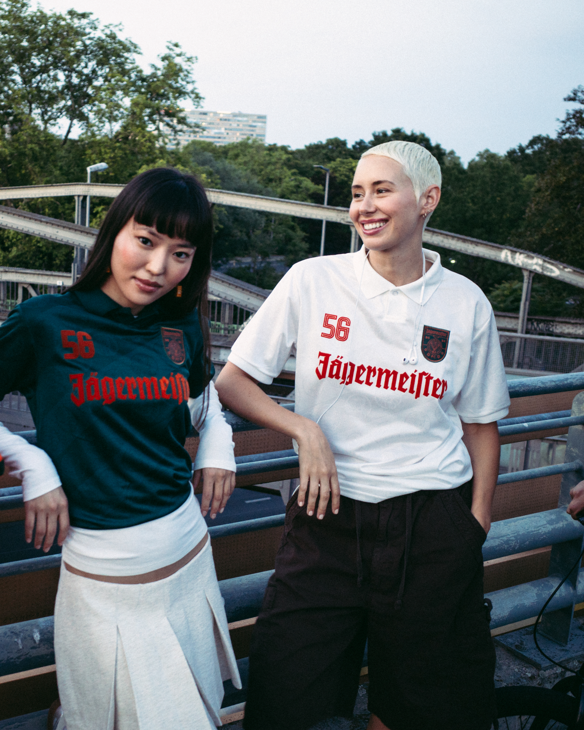 Patru persoane stând în aer liber, pe un pod sau o pasarelă, îmbrăcate în streetwear. O persoană poartă o jachetă portocalie intensă cu fermoar, alta poartă un tricou Jägermeister verde închis cu o fustă albă, a treia poartă un tricou alb Jägermeister cu pantaloni scurți închiși la culoare, iar a patra poartă un hanorac negru și o șapcă portocalie.