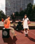 Scenă de lifestyle streetwear în aer liber, pe un teren de baschet, cu o clădire înaltă de apartamente în fundal. Un bărbat așezat într-un tricou Jägermeister portocaliu stă pe o boxă neagră, în timp ce o femeie în picioare, într-o rochie polo albă Jägermeister, ține o minge de baschet.
