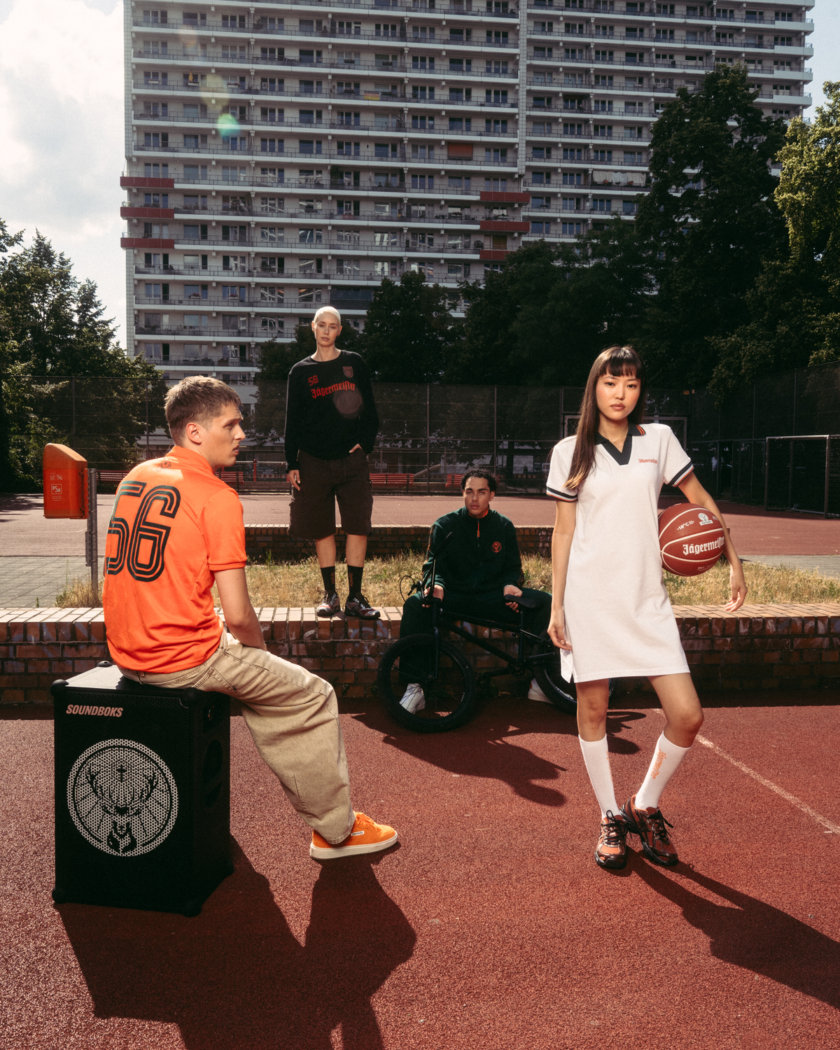 Scenă de lifestyle streetwear în aer liber, pe un teren de baschet, cu o clădire înaltă de apartamente în fundal. Un bărbat așezat într-un tricou Jägermeister portocaliu stă pe o boxă neagră, în timp ce o femeie în picioare, într-o rochie polo albă Jägermeister, ține o minge de baschet.