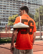 O persoană stă pe un teren sportiv în aer liber, purtând un tricou Jägermeister portocaliu intens cu un „56” mare imprimat pe spate. O pereche de adidași albi Jägermeister îi atârnă pe un umăr, iar în fundal se văd un gard din plasă metalică și blocuri de apartamente.