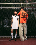 Două modele stau pe un teren de tenis roșu, în fața unui gard din plasă metalică. Modelul din stânga poartă o rochie polo sport albă Jägermeister cu borduri verde închis, șosete albe și pantofi negri.