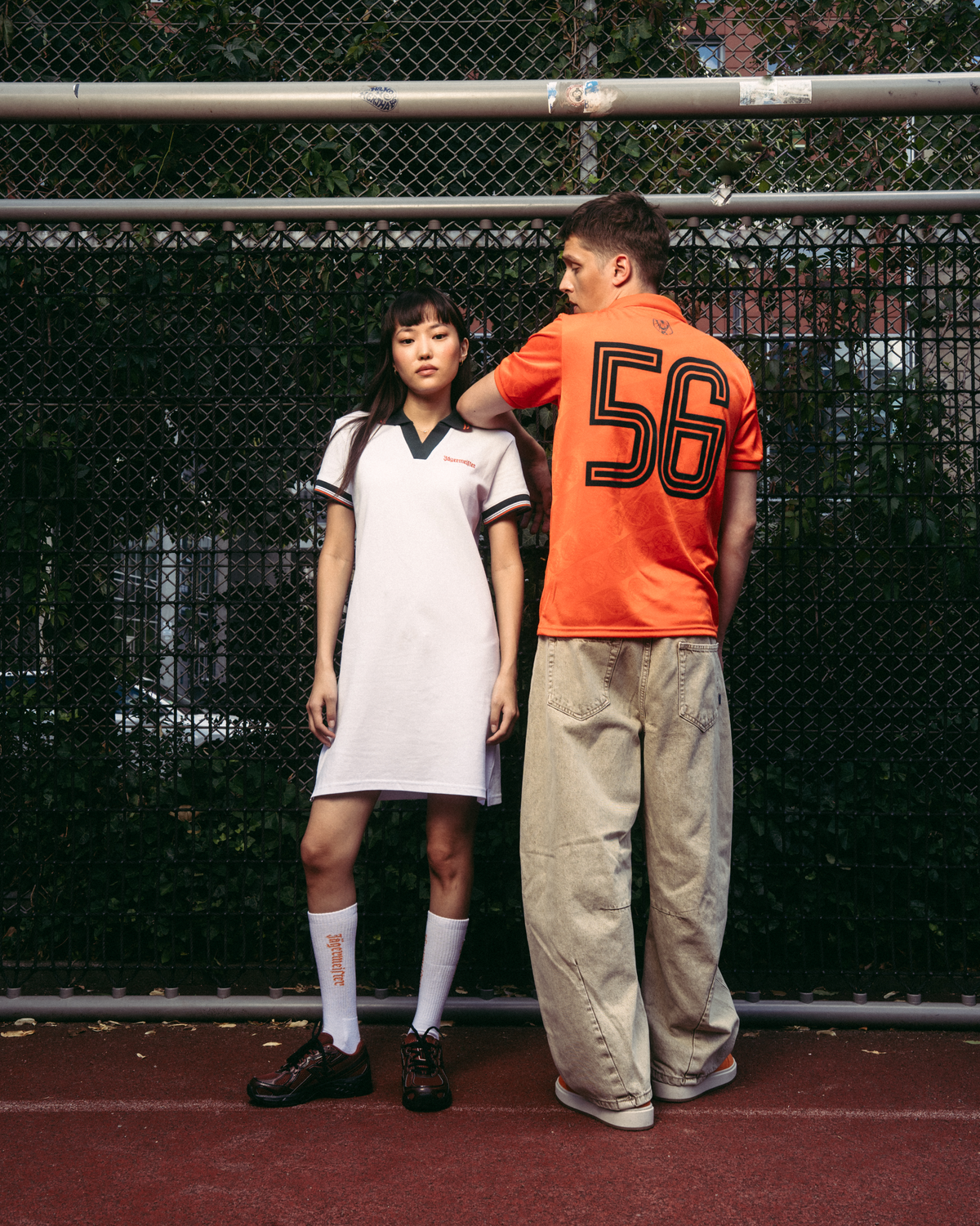 Două modele stau pe un teren de tenis roșu, în fața unui gard din plasă metalică. Modelul din stânga poartă o rochie polo sport albă Jägermeister cu borduri verde închis, șosete albe și pantofi negri.