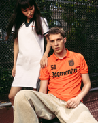 Un bărbat purtând un tricou Jägermeister portocaliu intens și pantaloni bej stă pe un teren, în timp ce o femeie într-o rochie polo albă Jägermeister stă în spatele lui, lângă un gard din plasă metalică.