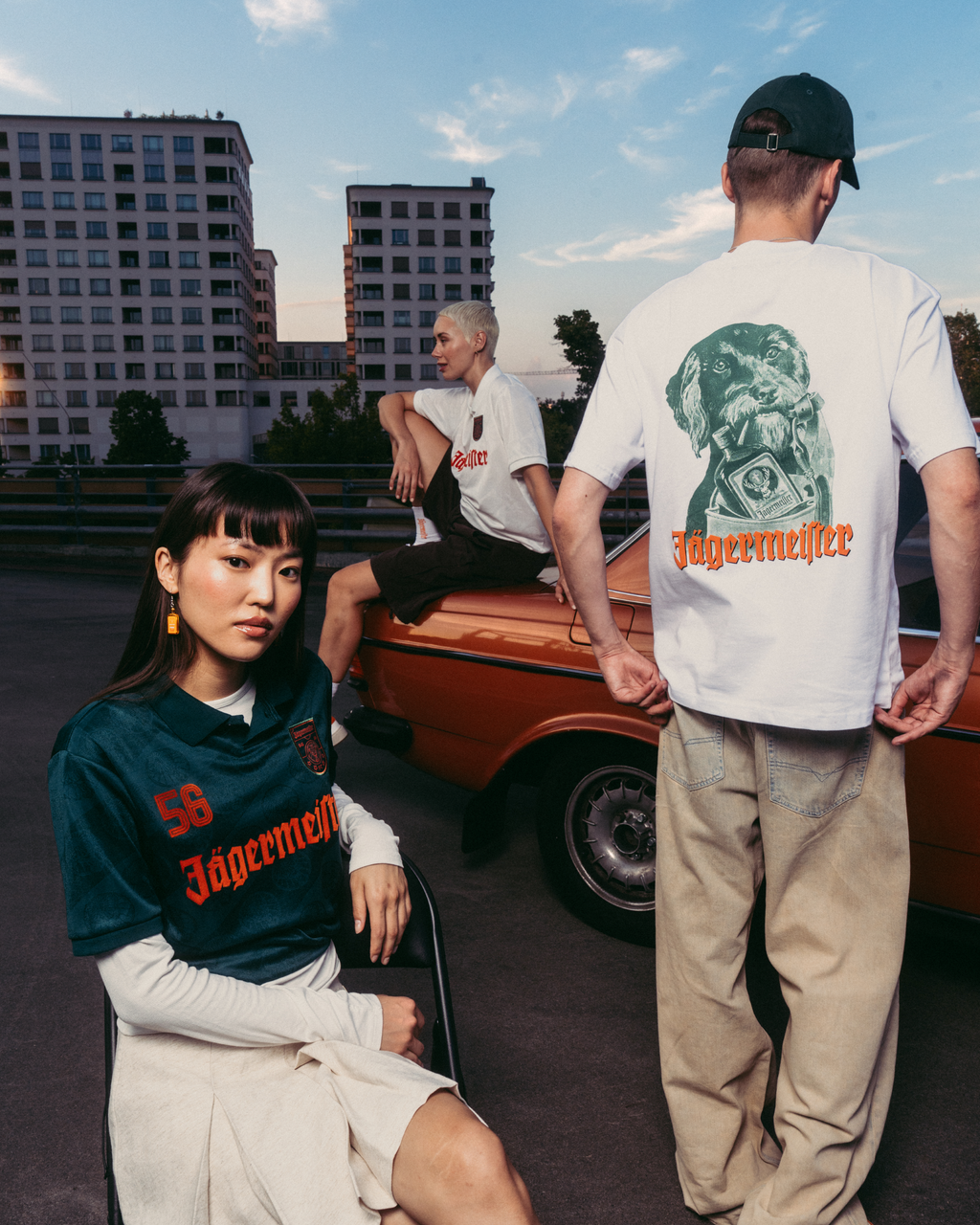 Trei modele purtând articole vestimentare cu brandul Jägermeister, într-un cadru urban în aer liber, cu o mașină vintage portocalie și blocuri de apartamente în fundal. Un model stă în prim-plan, purtând un tricou Jägermeister verde închis, altul stă cu spatele la cameră într-un tricou alb cu imprimeu grafic și pantaloni bej, iar al treilea model stă pe mașină într-un tricou alb.