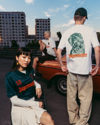 Trei modele purtând articole vestimentare cu brandul Jägermeister, într-un cadru urban în aer liber, cu o mașină vintage portocalie și blocuri de apartamente în fundal. Un model stă în prim-plan, purtând un tricou Jägermeister verde închis, altul stă cu spatele la cameră într-un tricou alb cu imprimeu grafic și pantaloni bej, iar al treilea model stă pe mașină într-un tricou alb.