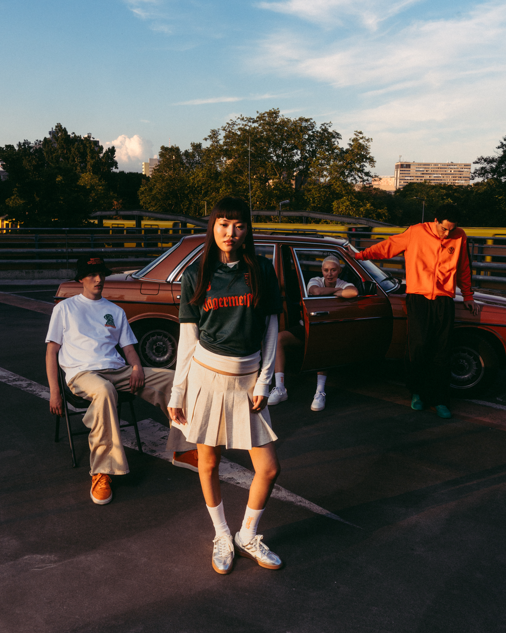 Un grup de patru tineri pozând în aer liber, pe un circuit, la apus, lângă o mașină vintage roșie. Figura centrală poartă un tricou Jäermeister verde închis cu lettering portocaliu, o fustă albă plisată și adidași albi.