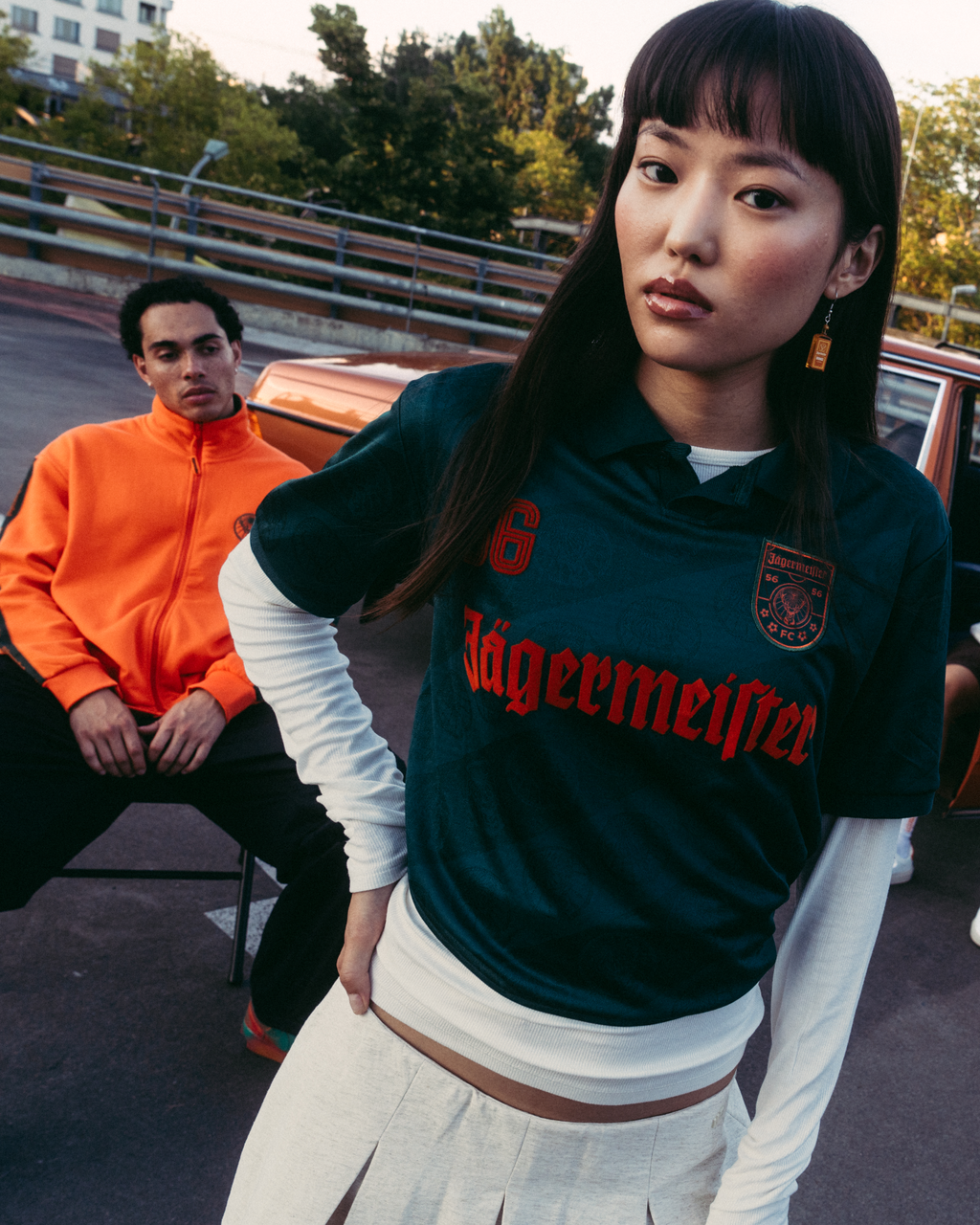 Un grup de patru persoane pozând în aer liber, în fața unei mașini vintage maro. Figura centrală poartă un tricou Jägermeister verde închis, iar ceilalți sunt îmbrăcați în streetwear casual.