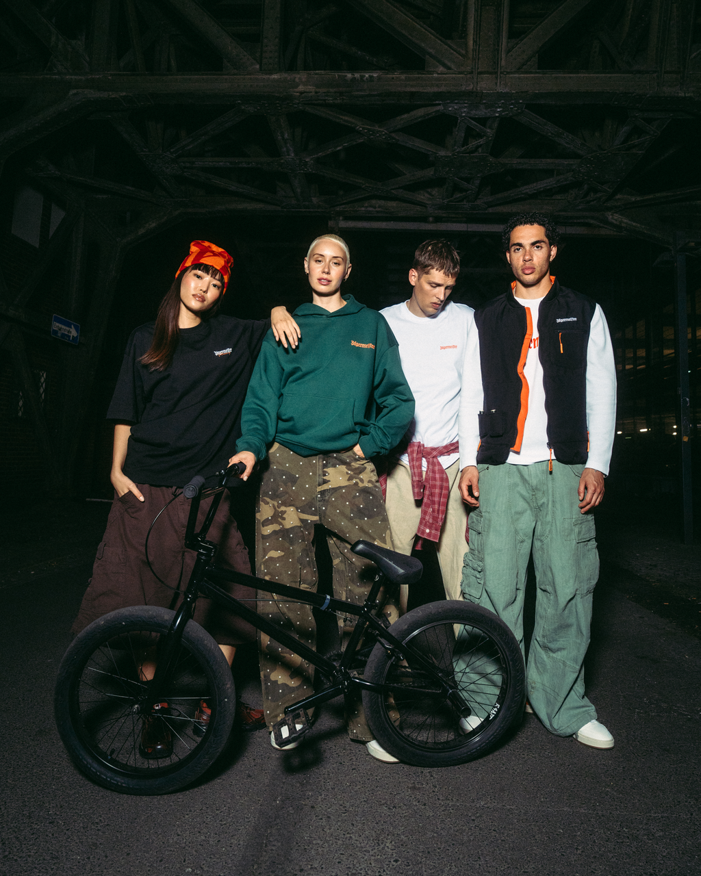 Patru tineri pozând cu o bicicletă BMX neagră, într-un decor întunecat, industrial, sub un pod, noaptea. Sunt îmbrăcați în ținute streetwear Jägermeister, inclusiv un hanorac verde, un tricou alb, o vestă neagră și pantaloni cargo.