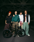 Patru tineri pozând cu o bicicletă BMX neagră, într-un decor întunecat, industrial, sub un pod, noaptea. Sunt îmbrăcați în ținute streetwear Jägermeister, inclusiv un hanorac verde, un tricou alb, o vestă neagră și pantaloni cargo.