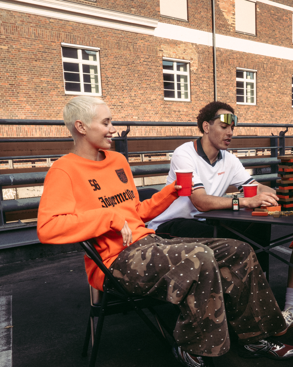 Patru persoane așezate în jurul unei mese mici pe o terasă pe acoperiș sau pe un pod, în aer liber, ținând pahare roșii. Sunt îmbrăcate în streetwear casual Jägermeister, inclusiv o bluză cu mânecă lungă portocalie și o cămașă polo albă.