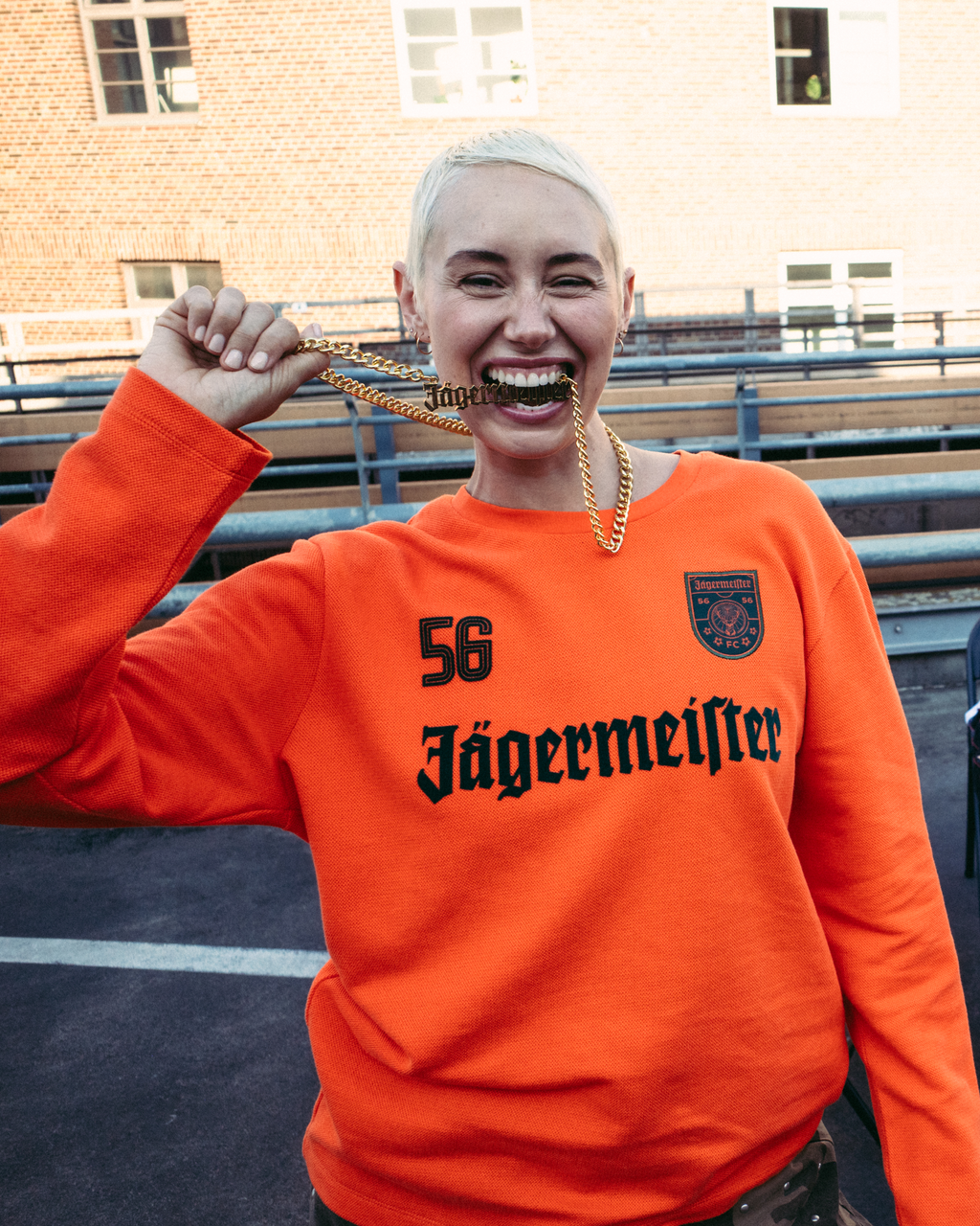 O femeie zâmbitoare, în prim-plan, poartă un hanorac portocaliu aprins Jägermeister cu „56 Jägermeister” imprimat cu negru pe față și ține un lanț auriu Jägermeister.
