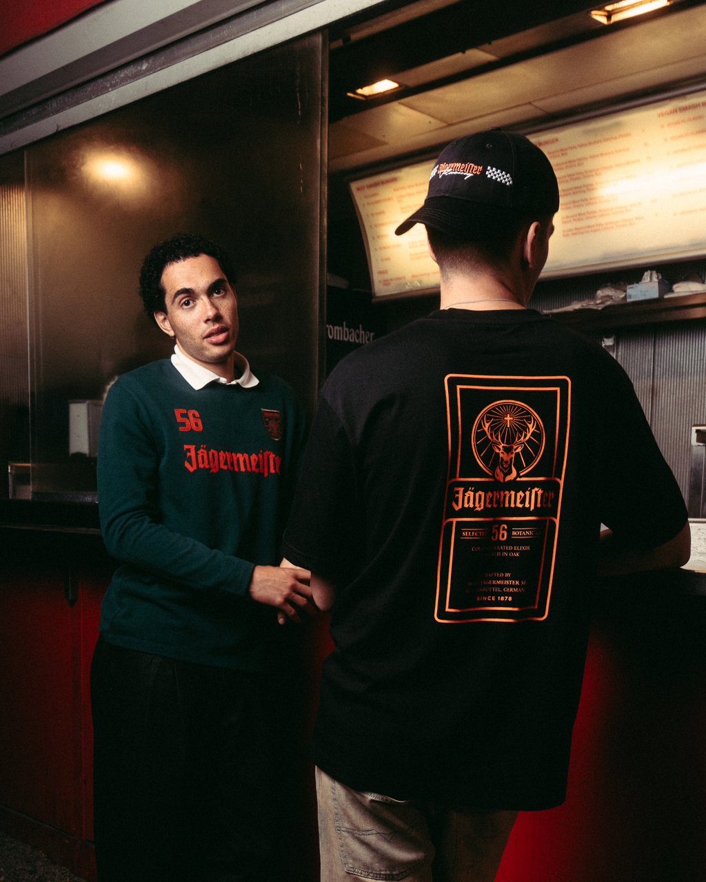 Doi bărbați stând afară, lângă un food truck roșu, noaptea. Unul dintre ei este cu fața la cameră și poartă o bluză cu mânecă lungă Jägermeister verde închis, cu text portocaliu, pantaloni negri și sneakers albi.