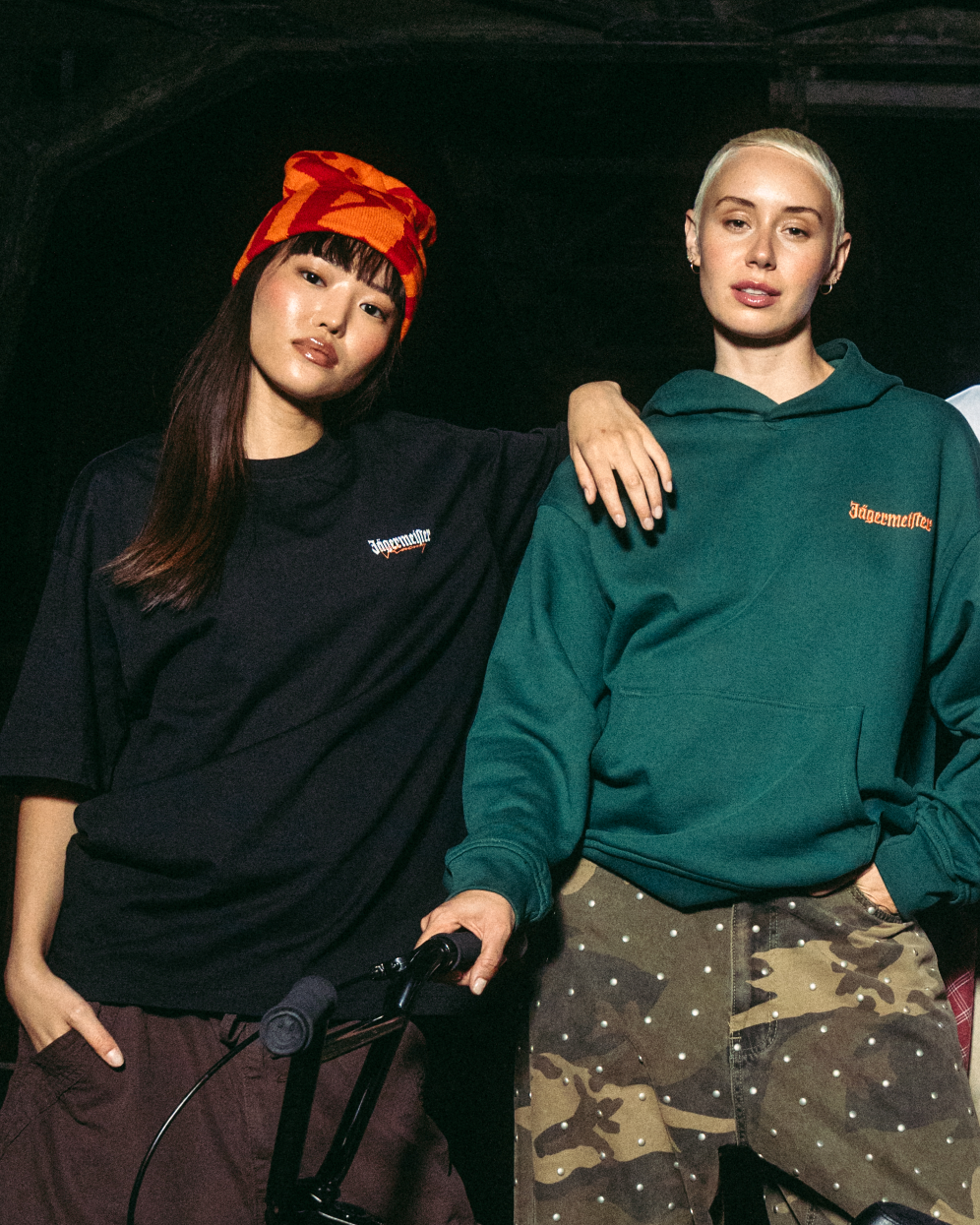 Two people wearing Jägermeister apparel pose indoors against a dark background. One person wears a black oversized T-shirt with a small Jägermeister logo and a red-orange patterned beanie, while the other wears a green Jägermeister hoodie with camouflage-patterned trousers. A bicycle handlebar is visible in the foreground, and the scene is lit with focused, moody lighting that highlights the clothing and branding.