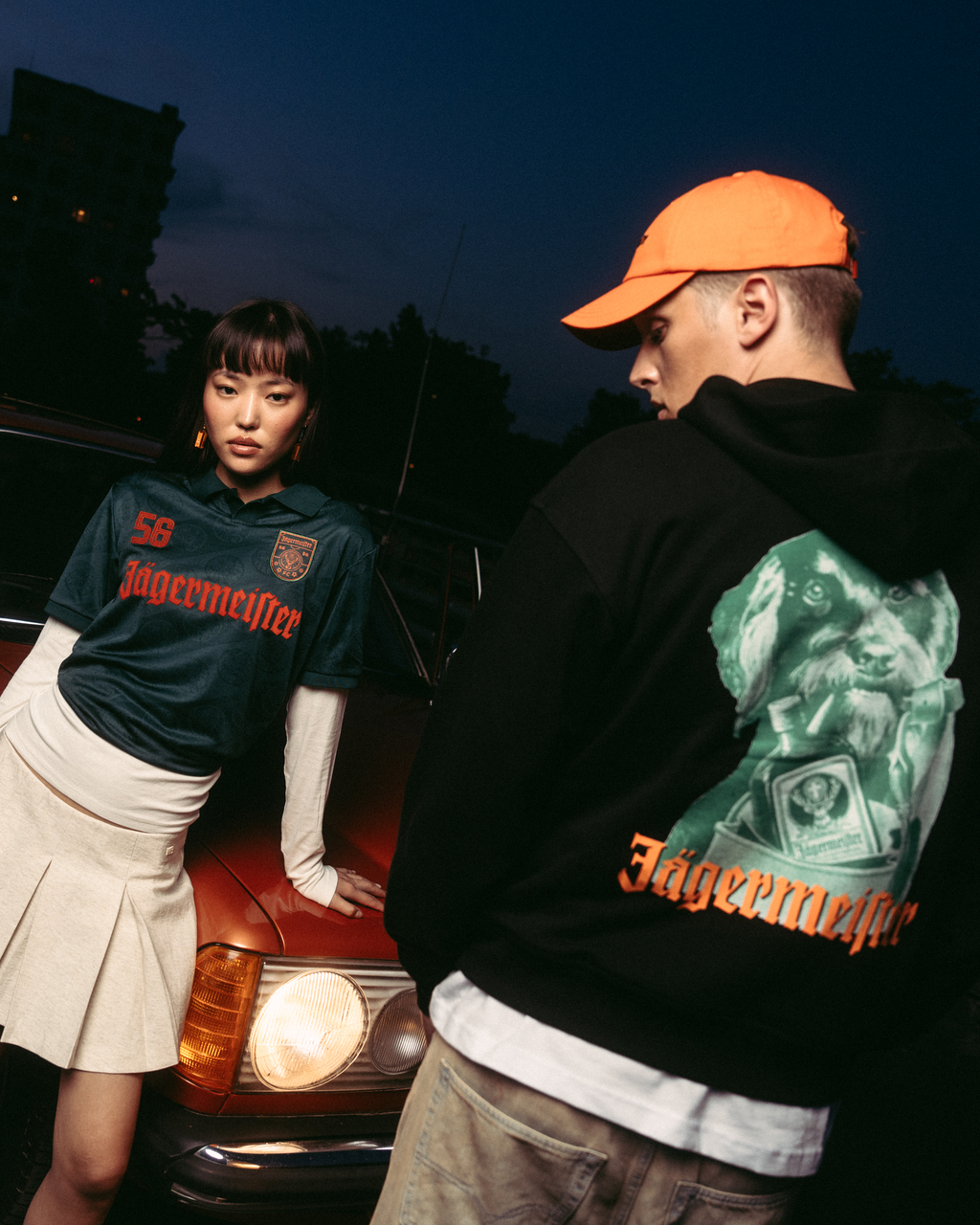 Two models posing in front of a vintage red car at dusk. One model wears a dark green 'Jägermeister' jersey with a white pleated skirt, and the other wears a black Jägermeister Waldi hoodie and an orange cap.