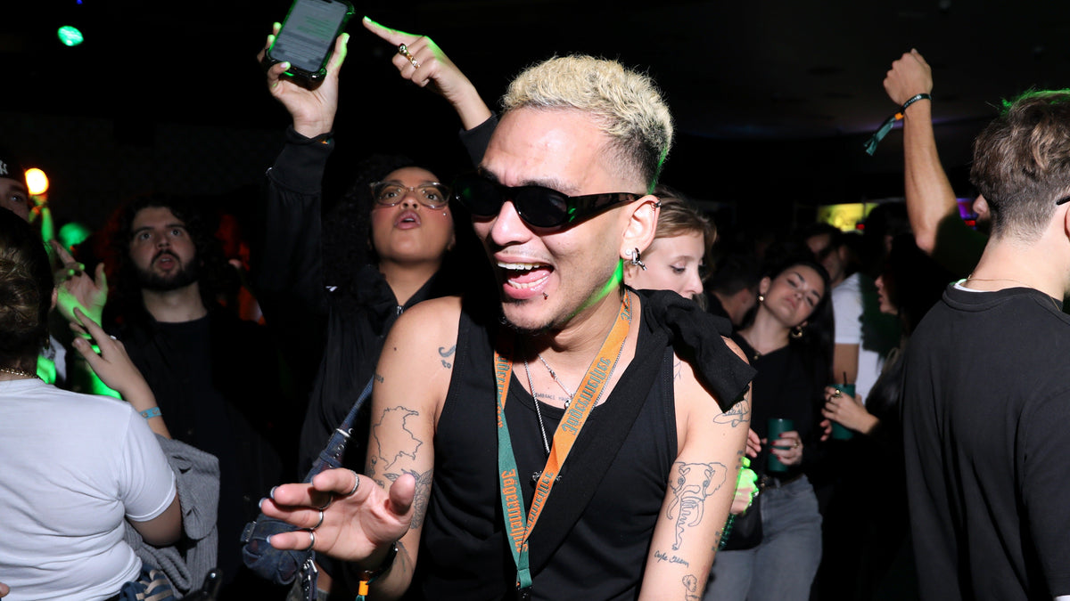 A person with short blonde hair, black sunglasses, and a black sleeveless top dancing in a crowded nightclub or event space with other people and green lighting visible.
