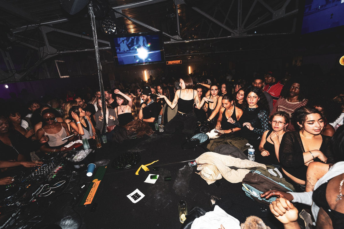 A crowded nightclub or party scene with many people dancing and sitting around a DJ booth under dim party lighting. A screen hangs overhead, and the environment is dark with black tables and stage lighting.