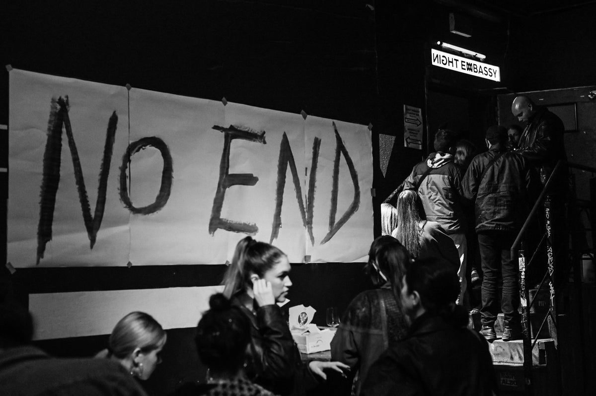 Black and white photograph of a crowd gathered around a large hand-painted 'NO END' banner, with people standing on a staircase in a dim indoor setting.
