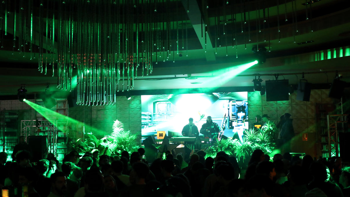 A live band performing on stage in a large venue, illuminated by dramatic green stage lighting and spotlights, with an audience in the foreground.