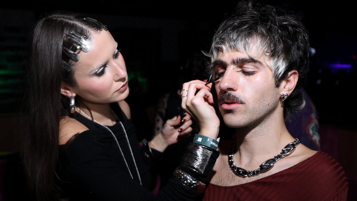 A woman with dramatic dark hair and silver clips applies makeup to a man with a mustache, earring, and chunky chain necklace in a dimly lit setting.