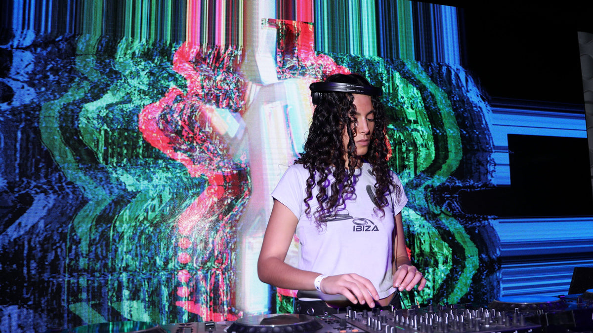 A DJ with long curly hair wearing a white t-shirt and headphones, mixing music behind a turntable in front of a vibrant, glitch-effect colored screen.