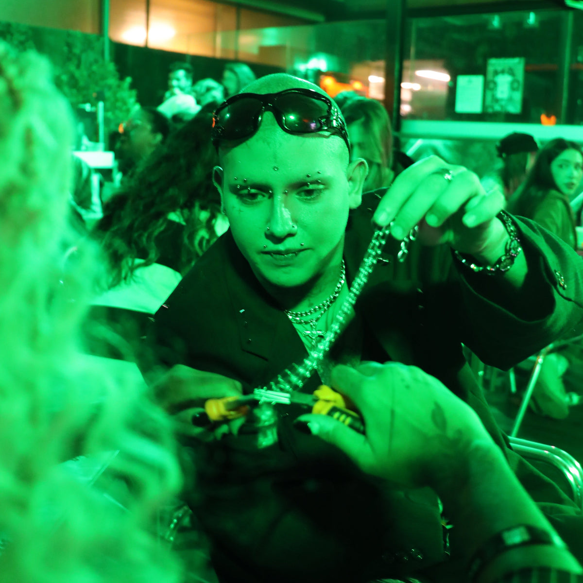 A person in a dark jacket and sunglasses holds a silver chain necklace in a crowded indoor setting, heavily tinted green with dramatic lighting.