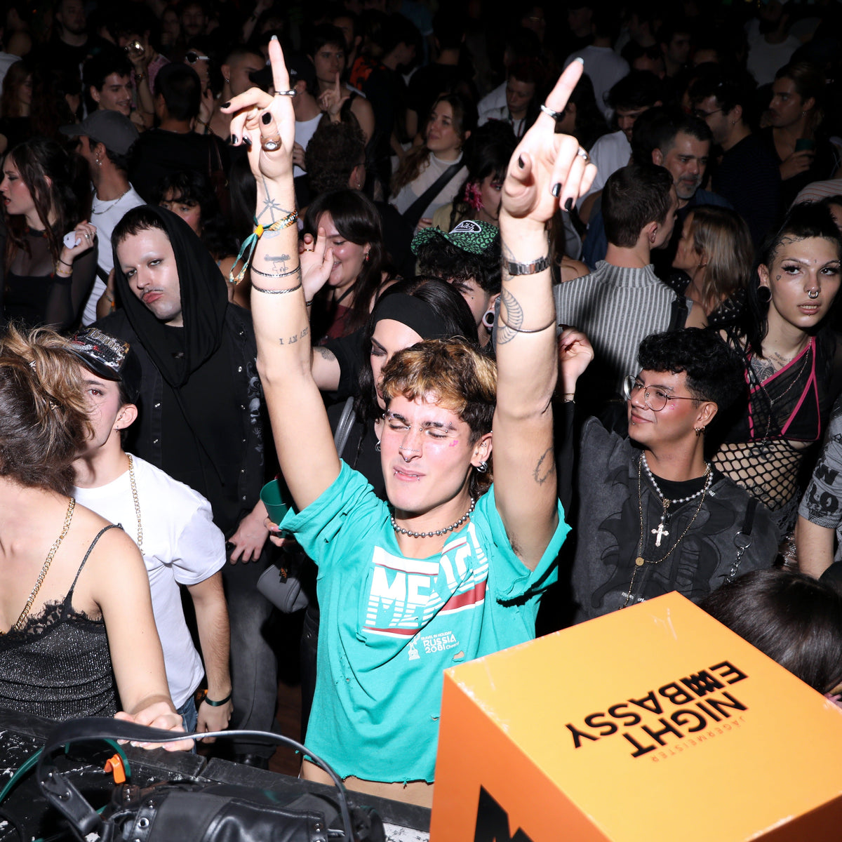 A crowd scene at a night event with a young man in the center wearing a bright teal graphic t-shirt and raising both hands in a gesture. In the foreground, an orange box with black text reading 'EMBASSY NIGHT' is partially visible.