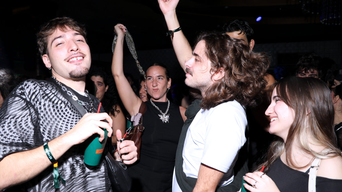 A group of young people dancing and celebrating in a dimly lit nightclub or party setting. The foreground features two men, one on the left wearing a black and white patterned shirt and holding a green drink, and another on the right with long brown hair in a white t-shirt.