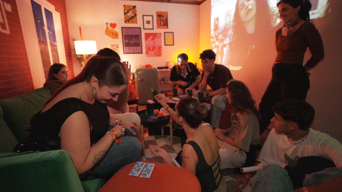 A group of young adults gathered in a dimly lit room, playing card games around a small table under warm ambient lighting and a projected scene on the wall.