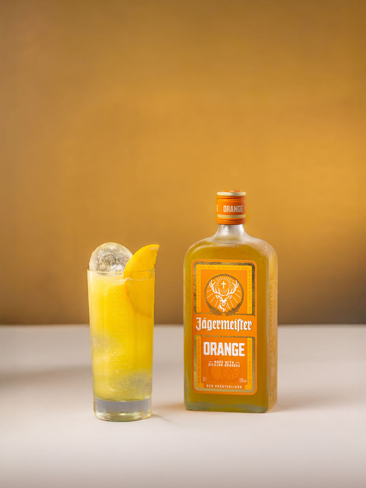 A bottle of Jägermeister Orange spirit and a tall glass filled with the same orange liquid, garnished with an orange slice and ice, set against a warm yellow studio background.