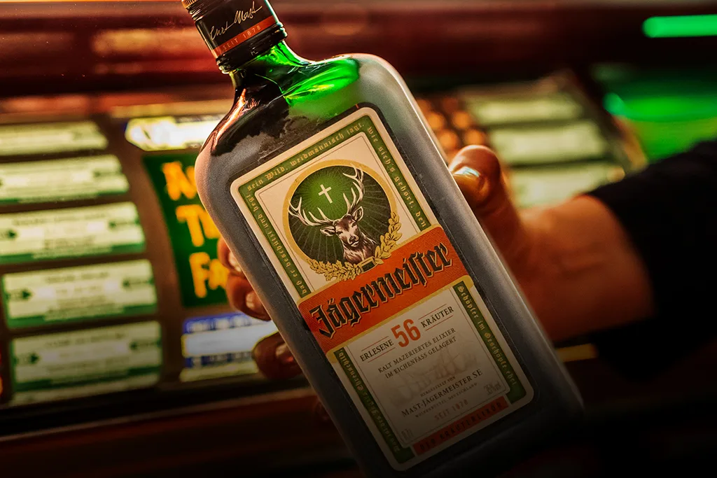 A hand holding a bottle of ice cold Jägermeister against a blurred bar background with green lights.