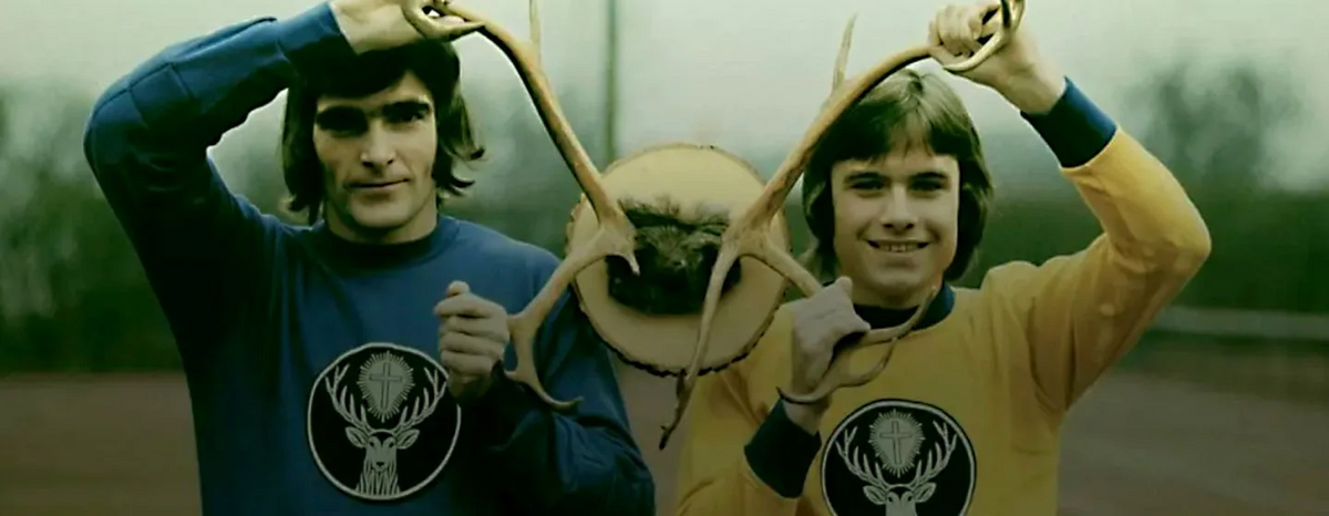 Two men outdoors holding deer antlers above their heads, wearing matching long-sleeve shirts with a black deer head logo. One shirt is blue and the other is yellow.