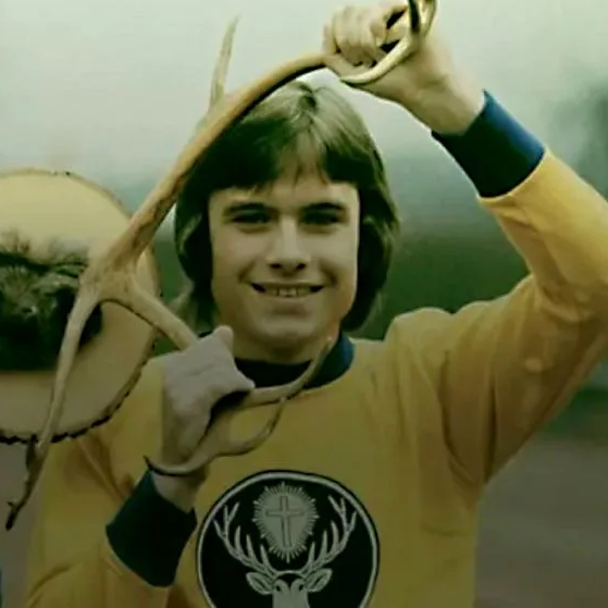 A men outdoors holding deer antlers above his head, wearing a yellow long-sleeve shirt with a black deer head logo.