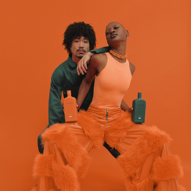 Two models pose with orange and green bottles against a solid orange background. The model in the foreground wears a peach bodysuit and voluminous orange faux-fur pants, while the model behind wears a dark green shirt and holds an orange bottle.