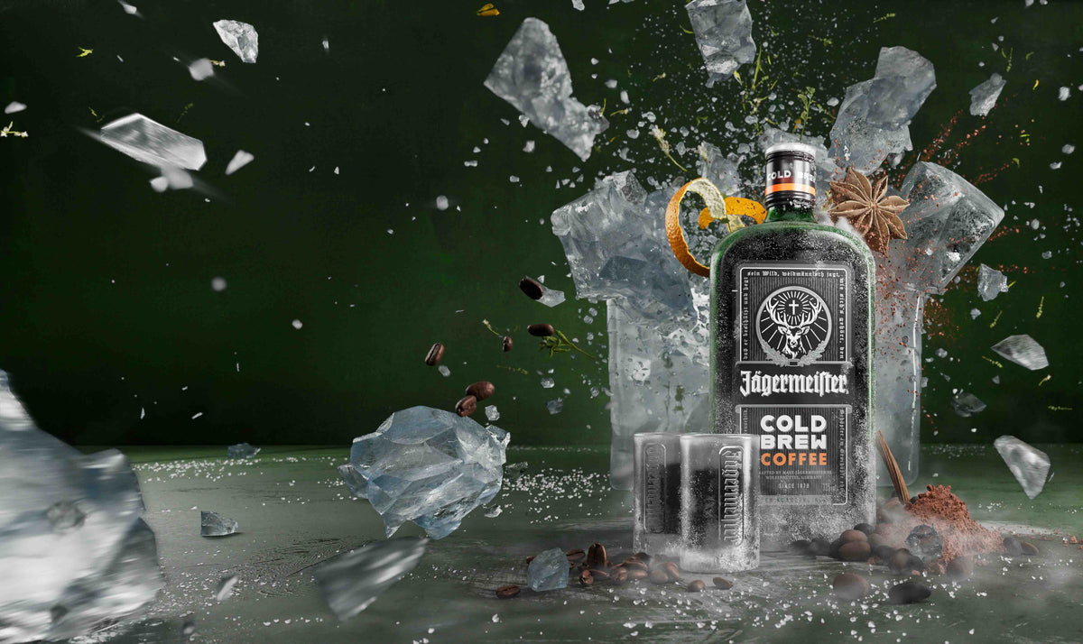 A bottle of Jägermeier Cold Brew Coffee garnished with an orange slice and star anise, surrounded by shattered ice, coffee beans, and two small glasses against a dark green background.