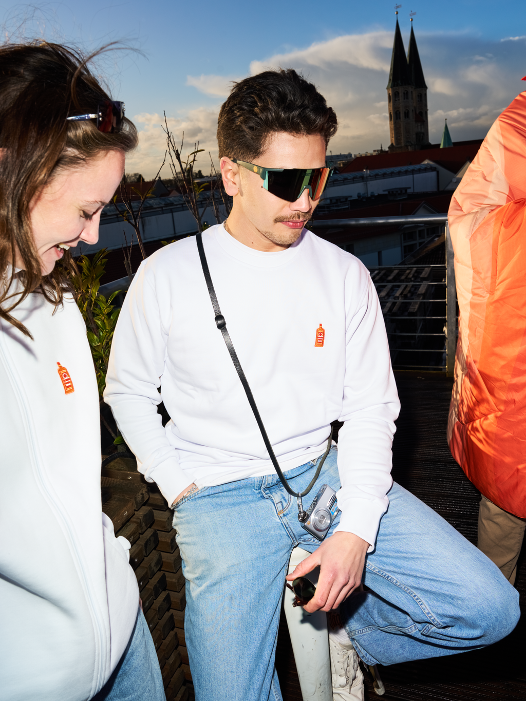 Two People wearing white Jägermeister sweatshirts and Jägermeister fast glasses on a rooftop terrace, with a church tower and city skyline in the background.