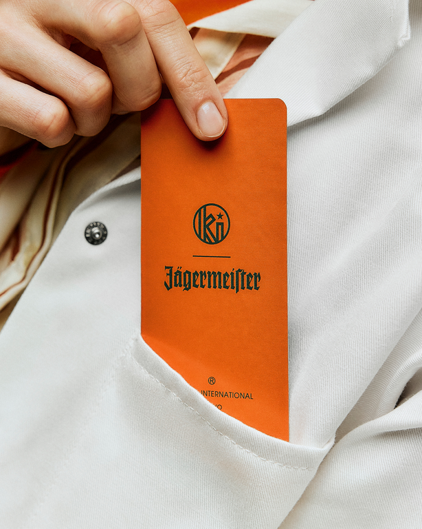 Etichetă Jägermeister și Kuumba pe o hârtie de parfum portocalie, ținută în mână pe un fundal de halat alb