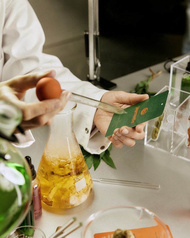 Persoană care ține o sticlă cu lichid galben și o hârtie de parfum verde, înconjurată de obiecte de laborator.